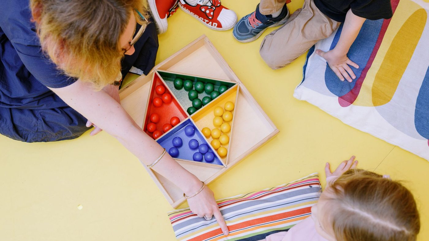 Une adulte présente à des enfants assis par terre un plateau de perles à trier par couleur