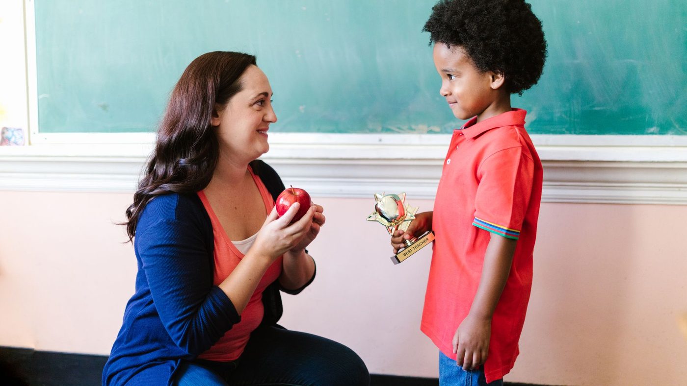 Une maîtresse assise montre une pomme à un petit garçon, devant le tableau de la classe