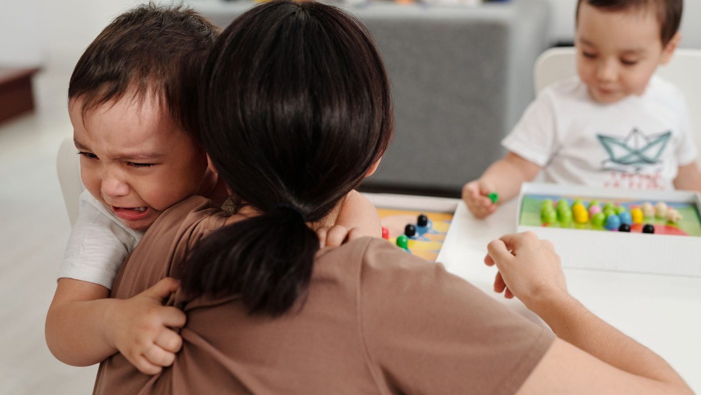 Une mère serre contre elle un petit garçon qui pleure, pendant qu'un autre enfant joue à la table