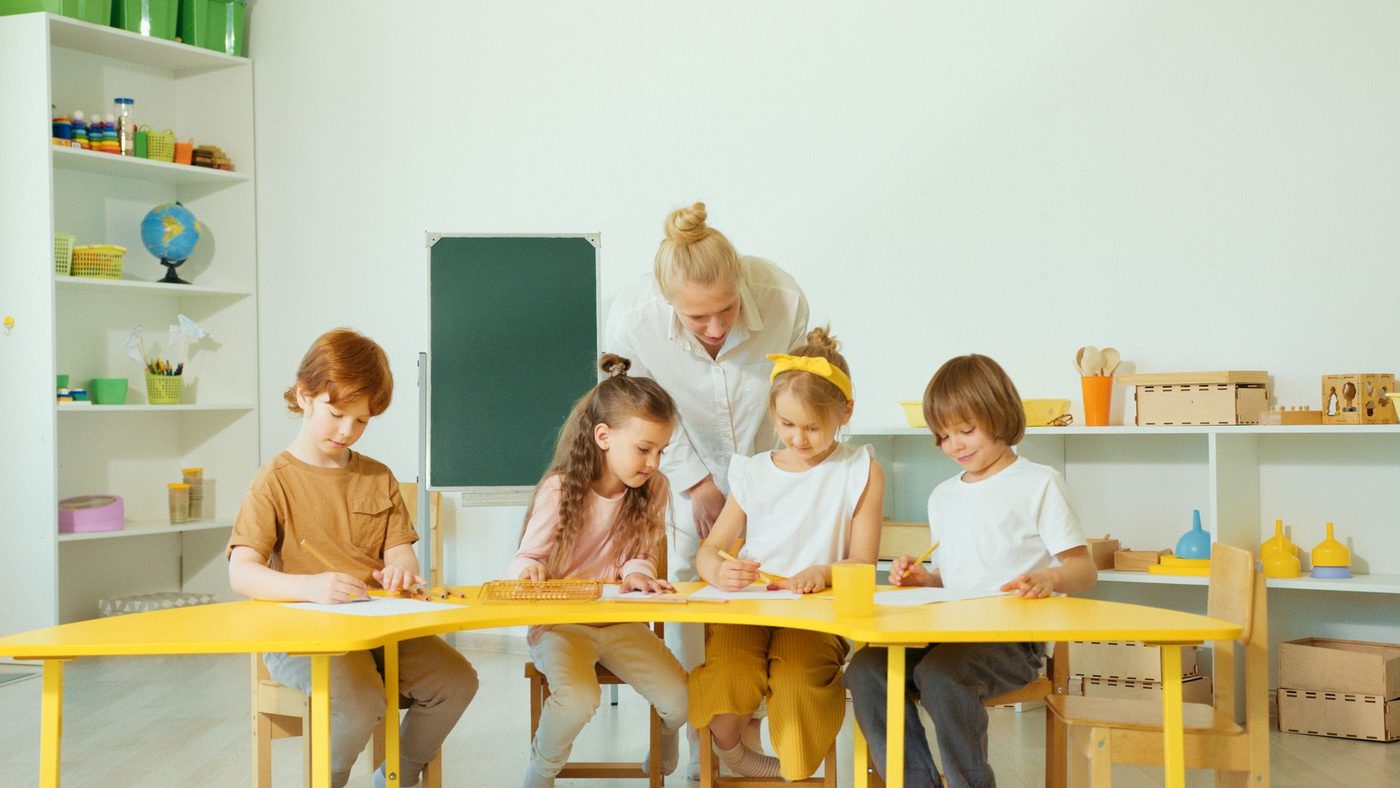 Une maîtresse penchée sur une table jaune où travaillent quatre enfants, devant des étagères basses