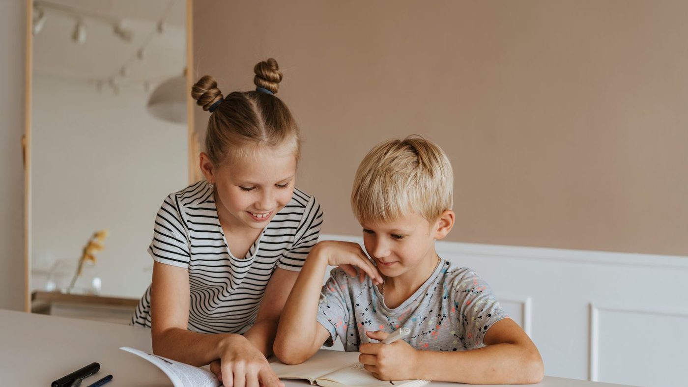 Une enfant plus grande montre un livre à un plus jeune, assis côte à côte à une table