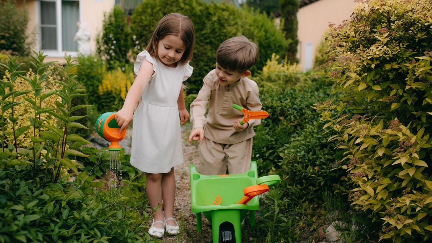 Deux jeunes enfants jardinent dans une allée, l'un avec un arrosoir, l'autre avec une petite brouette verte
