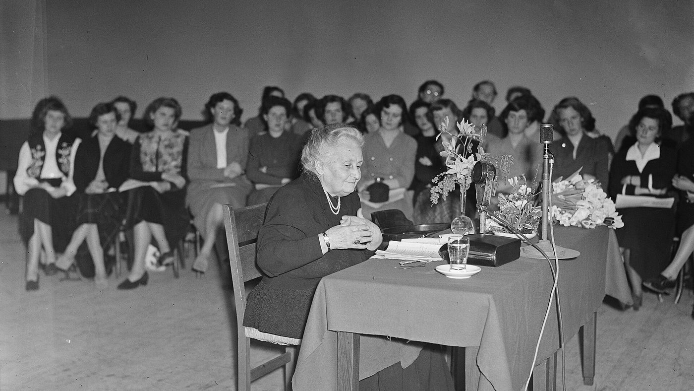 Maria Montessori assise à une table devant un micro, face à un public, lors d'une conférence à Amsterdam en 1950