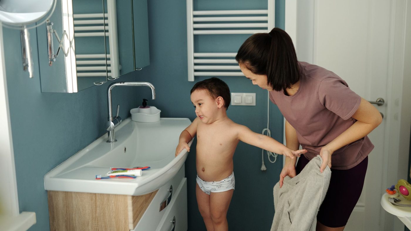 Un petit garçon en culotte devant le lavabo de la salle de bains, sa mère à côté de lui avec une serviette